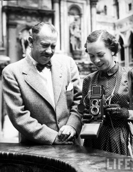 American novelist John Steinbeck standing beside wife, writer Elaine Andersen, who is using Rolleiflex camera around neck